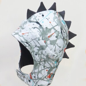 Op de afbeelding: Een witte stoffen hoofdbedekking met een grijs, turquoise en oranje spetterverf ontwerp. De hoofdbedekking heeft een zwart interieur en een rij zwarte driehoekige spikes langs de bovenrand.
