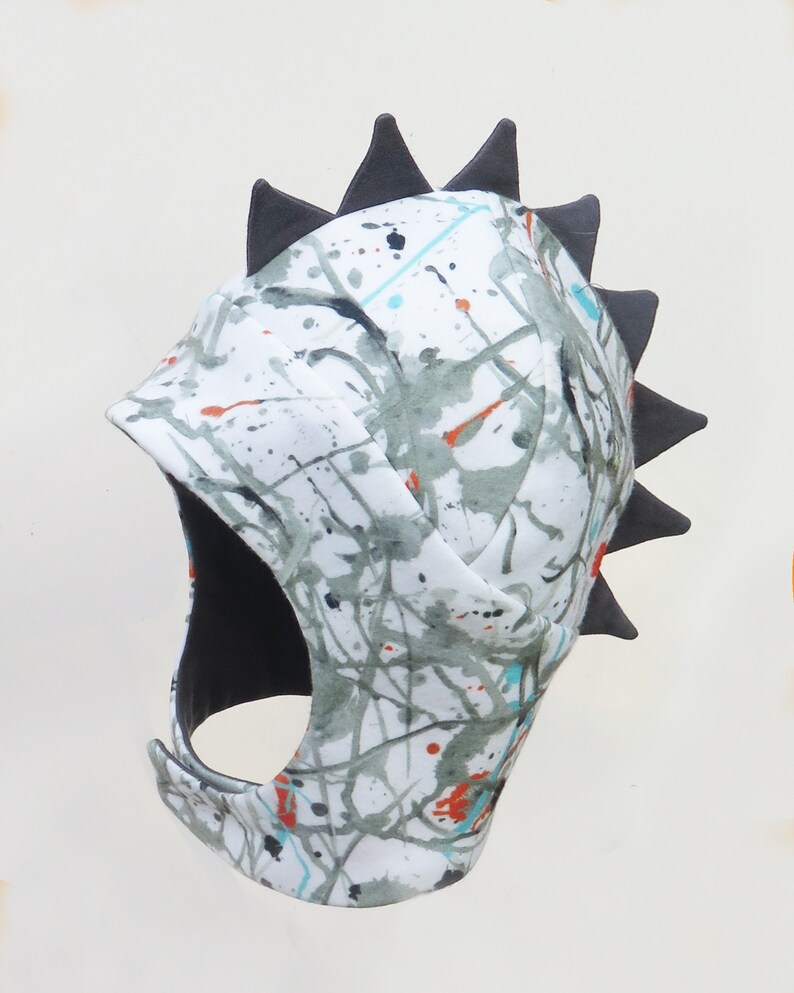 Op de afbeelding: Een witte stoffen hoofdbedekking met een grijs, turquoise en oranje spetterverf ontwerp. De hoofdbedekking heeft een zwart interieur en een rij zwarte driehoekige spikes langs de bovenrand.