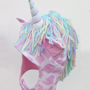 Op de afbeelding: Een roze eenhoornhoed met een witte hoorn en pastelkleurige garen manen. De hoed heeft een roze en wit giraffenprint patroon en roze oordetails. De hoorn is wit met pastelblauwe, groene en paarse strepen.