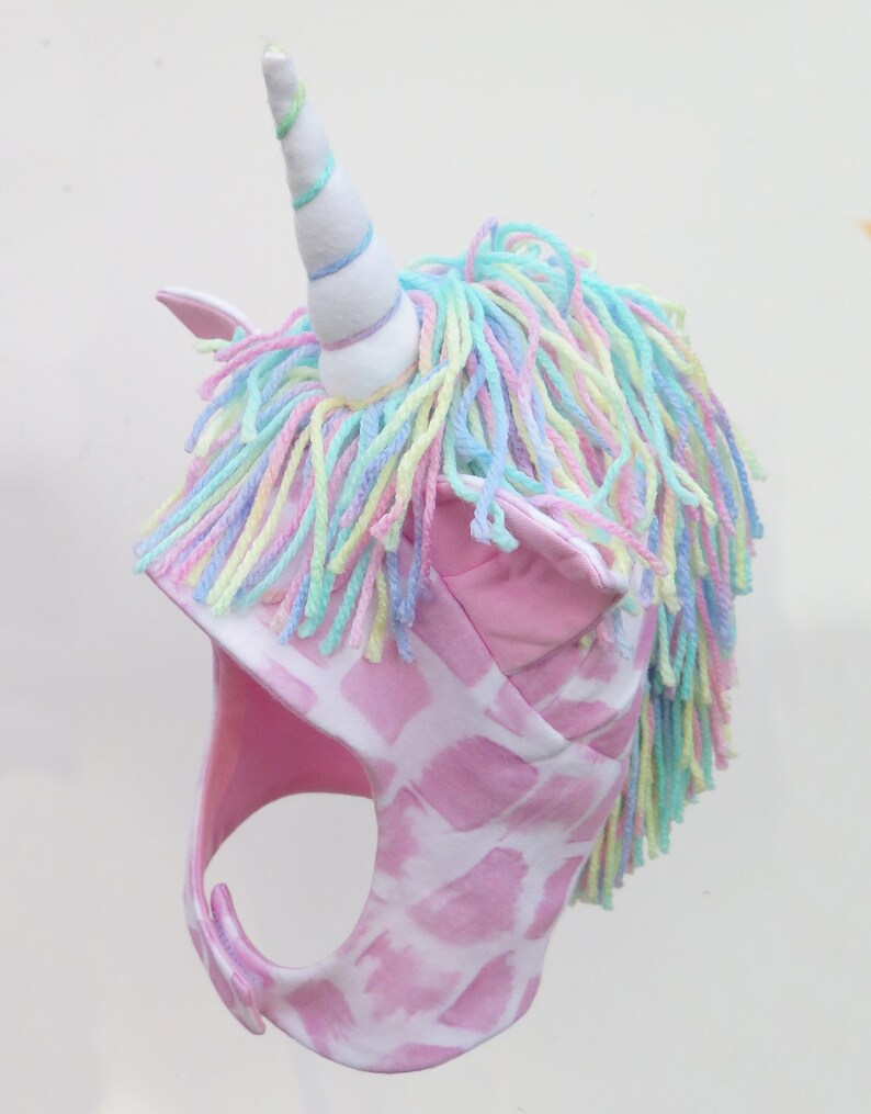 Op de afbeelding: Een roze eenhoornhoed met een witte hoorn en pastelkleurige garen manen. De hoed heeft een roze en wit giraffenprint patroon en roze oordetails. De hoorn is wit met pastelblauwe, groene en paarse strepen.