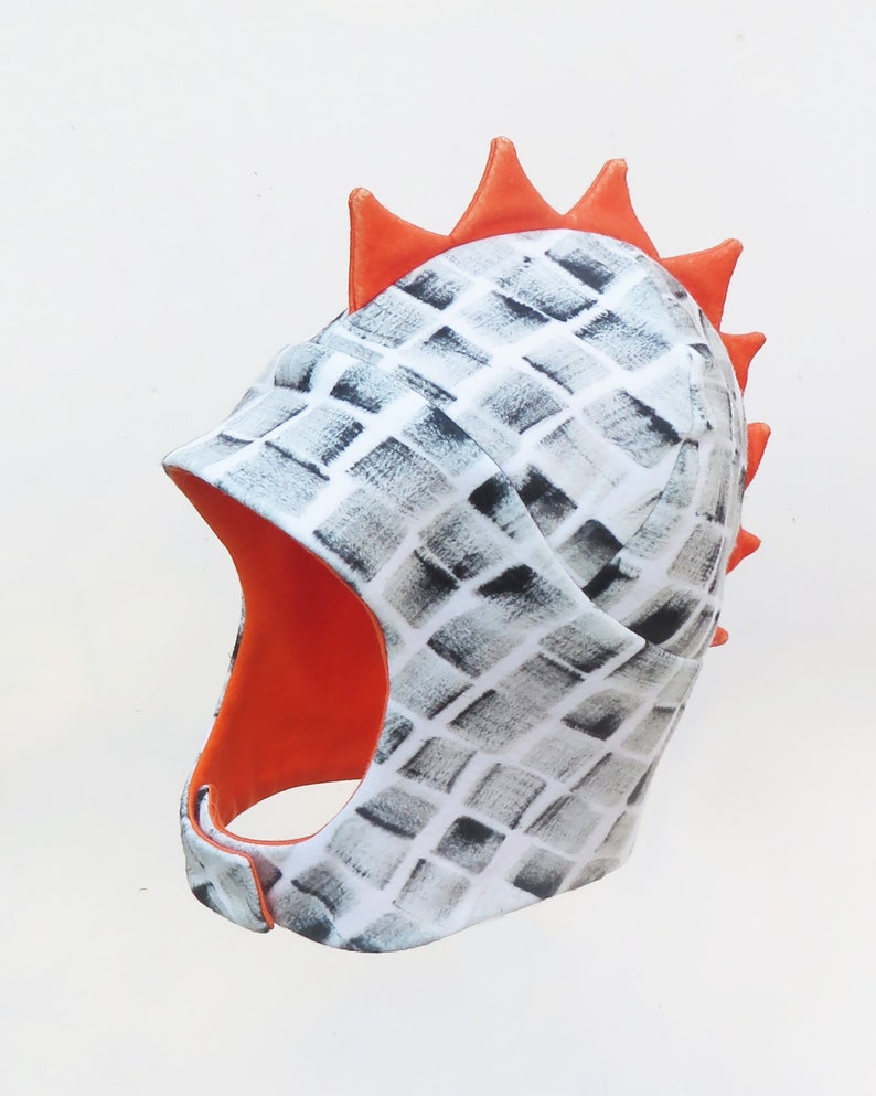 Op de afbeelding: Een unieke helmvormige hoed met een wit en grijs ruitpatroon. De binnenkant en de decoratieve stekels aan de achterkant zijn fel oranje. De hoed heeft een kinband en bedekt het hoofd en de nek.