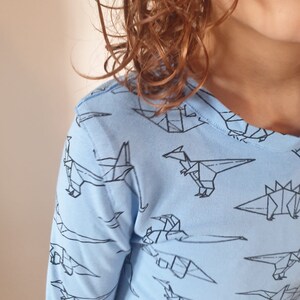 Puede incluir: Una camisa de manga larga azul claro con un patrón de dinosaurios de origami negros.