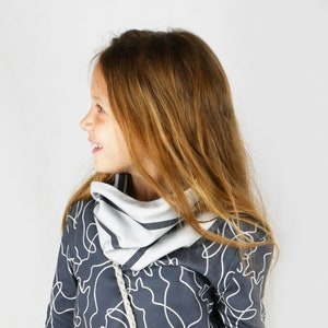 Può includere: Una ragazza giovane con lunghi capelli castani indossa una felpa con cappuccio grigio scuro con una sciarpa a motivi bianchi e grigi. La felpa ha un design astratto bianco.