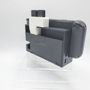 May include: A gray Nintendo Switch dock with a white cartridge holder attached. Two game cartridges are inserted into the holder. The dock is on a clear acrylic stand.