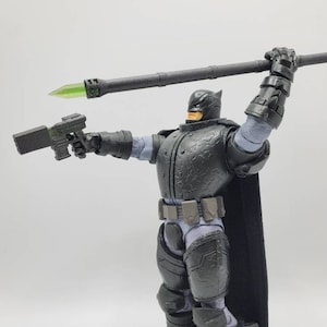 May include: A black and grey action figure of Batman wearing a black cape and holding a black and green weapon. The figure is posed with one arm raised, holding a long black weapon with a green tip.