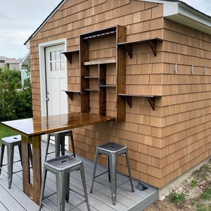 May include: A wooden folding table with a brown top and a dark brown base, attached to a wall with a built-in shelf unit. Four metal stools are placed around the table on a gray composite deck.