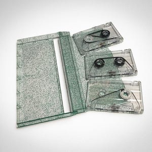 Può includere: Un astuccio per musicassette in plastica trasparente e tre musicassette. L'astuccio è aperto, con un design verde maculato. Le musicassette sono trasparenti, rivelando i meccanismi interni e il nastro nero.