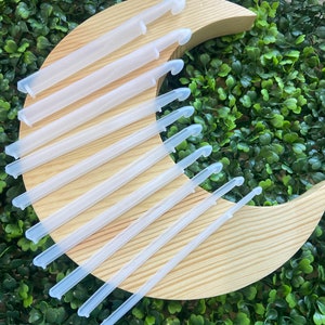 May include: A collection of clear plastic crochet hooks arranged on a light brown wooden crescent moon shape. The hooks are evenly spaced and angled, with a curved hook end and a straight handle. The background is a dense green hedge.