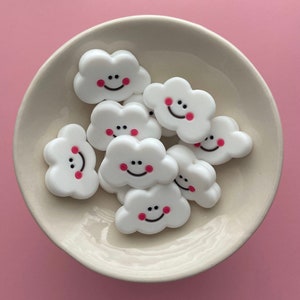 May include: A collection of white cloud-shaped charms with pink cheeks and black smiling faces. The charms are arranged in a bowl.