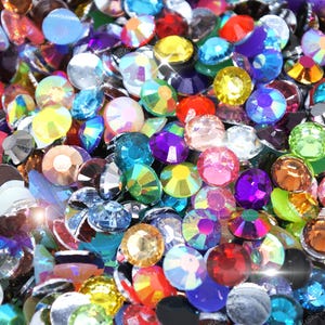 May include: A close-up of a collection of multi-coloured rhinestones. The gems are in various shapes and sizes, with colours including red, blue, yellow, purple, and clear. Light reflects off the facets, creating a sparkling effect.