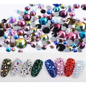 May include: Assortment of colourful rhinestones in various shapes and sizes. The rhinestones are scattered on a white surface. There are six nail art designs featuring different colours and patterns of rhinestones.