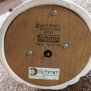 May include: The underside of a vintage Schmid music box. The wooden base features the text "Walt Disney Productions, Japan" and "Schmid Hand Painted." A sticker reads "203 Mickey Mouse Club March." The box is white with a wooden base.
