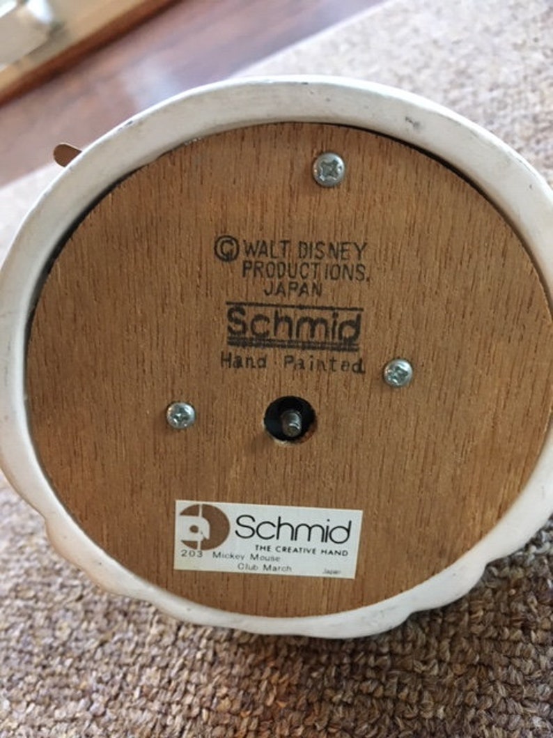 May include: The underside of a vintage Schmid music box. The wooden base features the text "Walt Disney Productions, Japan" and "Schmid Hand Painted." A sticker reads "203 Mickey Mouse Club March." The box is white with a wooden base.