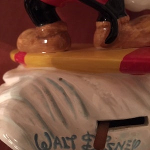 May include: A vintage Mickey Mouse figurine, featuring red shorts, black legs, and brown shoes. The figurine is standing on a yellow and red surfboard, atop a white base with the text "Walt Disney Characters" in blue.