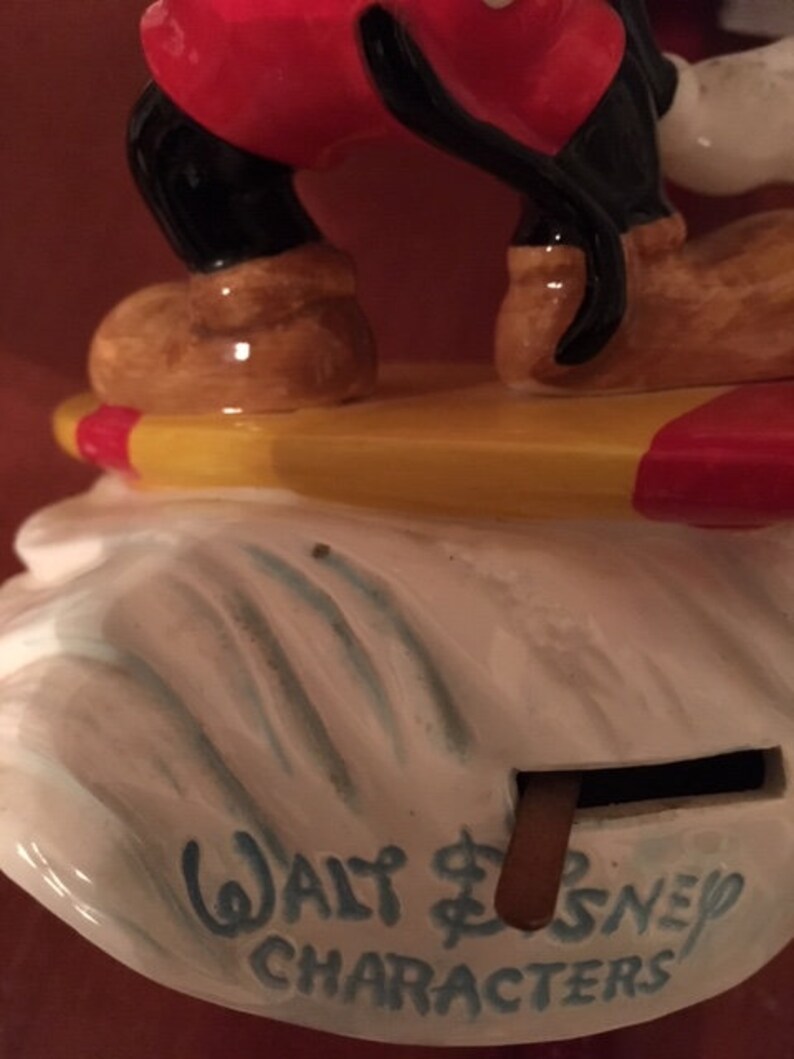 May include: A vintage Mickey Mouse figurine, featuring red shorts, black legs, and brown shoes. The figurine is standing on a yellow and red surfboard, atop a white base with the text "Walt Disney Characters" in blue.