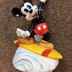 May include: A ceramic figurine of Mickey Mouse surfing. Mickey is wearing red shorts and brown shoes, and is standing on a yellow surfboard with red accents. The base is a white wave design.