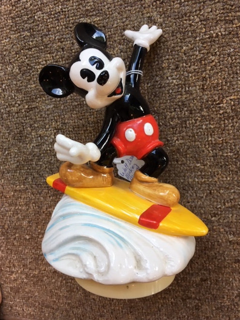May include: A ceramic figurine of Mickey Mouse surfing. Mickey is wearing red shorts and brown shoes, and is standing on a yellow surfboard with red accents. The base is a white wave design.