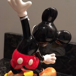 May include: A ceramic figurine of Mickey Mouse in a yoga pose. The figure is predominantly black, red, and white, with tan shoes and a yellow base with red stripes. The figurine has a glossy finish and is a collectible item.