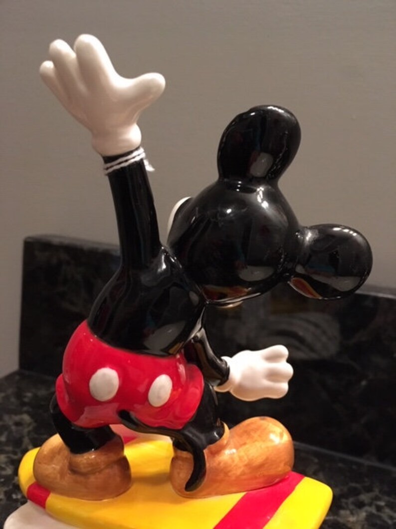 May include: A ceramic figurine of Mickey Mouse in a yoga pose. The figure is predominantly black, red, and white, with tan shoes and a yellow base with red stripes. The figurine has a glossy finish and is a collectible item.