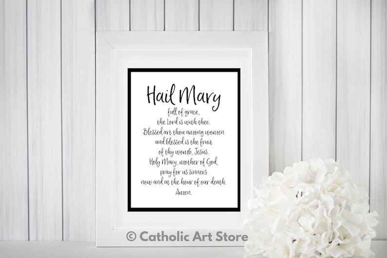 May include: A black and white printable wall art print with the text "Hail Mary" and the Hail Mary prayer. The print is framed in a white frame and is sitting on a white wood surface.
