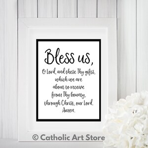 May include: A white framed print with black text that reads "Bless us, O Lord, and these Thy gifts, which we are about to receive from Thy bounty, through Christ, our Lord. Amen." The print is on a white wood background with a white flower in the bottom right corner. The text "Catholic Art Store" is in the bottom center of the image.