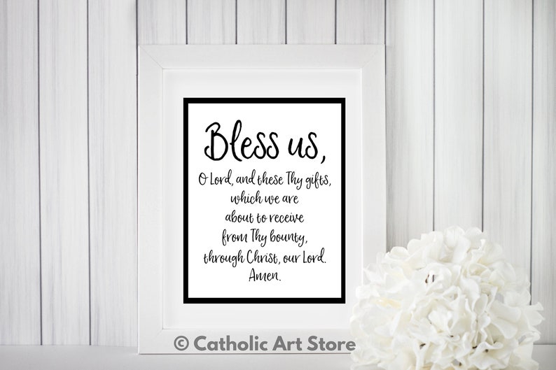 May include: A white framed print with black text that reads "Bless us, O Lord, and these Thy gifts, which we are about to receive from Thy bounty, through Christ, our Lord. Amen." The print is on a white wood background with a white flower in the bottom right corner. The text "Catholic Art Store" is in the bottom center of the image.