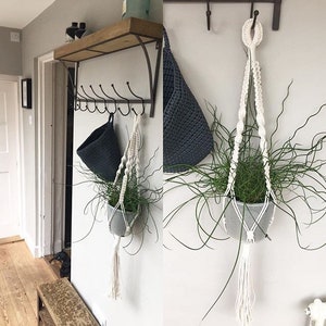 May include: Two macrame plant hangers with white cords and tassels hanging from a wall with a wooden shelf and black metal hooks. The hangers are holding small potted plants.