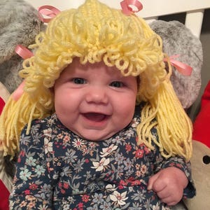 May include: A baby wearing a yellow yarn wig with pink ribbons, a floral print dress, and a big smile.