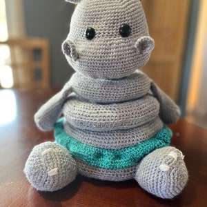 May include: A grey crocheted hippopotamus toy with black eyes and a teal skirt. The hippo is sitting on a wooden surface.
