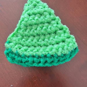 May include: A handmade, crocheted item in the shape of a watermelon slice. The top portion is light green, and the bottom is a darker green. The item is made of a textured yarn, and the stitches are visible.