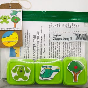 Puede incluir: Una bolsa verde con cremallera que contiene tres contenedores cuadrados verdes. Cada contenedor tiene una imagen en la parte delantera: un perro verde, un helicóptero verde y un árbol verde con una manzana roja. La bolsa está etiquetada como "Zippa Bag S" y tiene una pegatina amarilla con el texto "Toys for Talking".