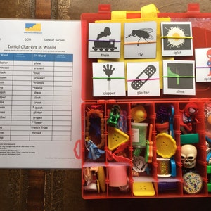 May include: A red plastic storage box containing a variety of small toys and objects. The box has a clear lid and is labelled "Initial Clusters in Words". The lid features a chart with a list of words and their corresponding initial consonant clusters. There are also several cards with pictures and words on them, such as "train", "fly", "splat", "clapper", "plaster", "slime", and "spiderman".
