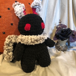 May include: Black crocheted plush toy with red cheeks and gray fur ears and scarf. The toy is sitting on a white blanket.