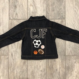 May include: A dark blue denim jacket with the letters "CJF" embroidered on the back in white. The jacket has four sports-themed patches on the back: a football, a baseball, a rugby ball, and a basketball.