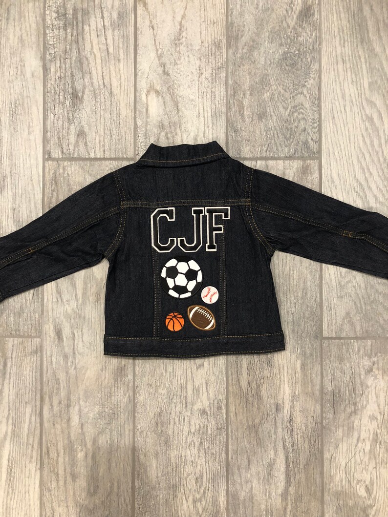May include: A dark blue denim jacket with the letters "CJF" embroidered on the back in white. The jacket has four sports-themed patches on the back: a football, a baseball, a rugby ball, and a basketball.