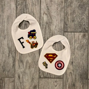 May include: Two white baby bibs with embroidered designs. One bib has a train, a bulldozer, and the letter "F". The other bib has a Superman logo, a lightning bolt, and a red, white, and blue Captain America logo.