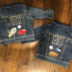 May include: Two denim jackets with personalised patches. One jacket has the name "JAY" and patches of a rocket, a baseball, and a dump truck. The other jacket has the name "MO" and patches of a baseball, a planet, and a dump truck.