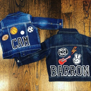 May include: Two denim jackets with personalised patches. One jacket has a basketball, baseball, football, soccer ball, and the name "CAM". The other jacket has a "Rock 'n' Roll" patch, a lightning bolt, a peace sign, a red electric guitar, and the name "BARRON".