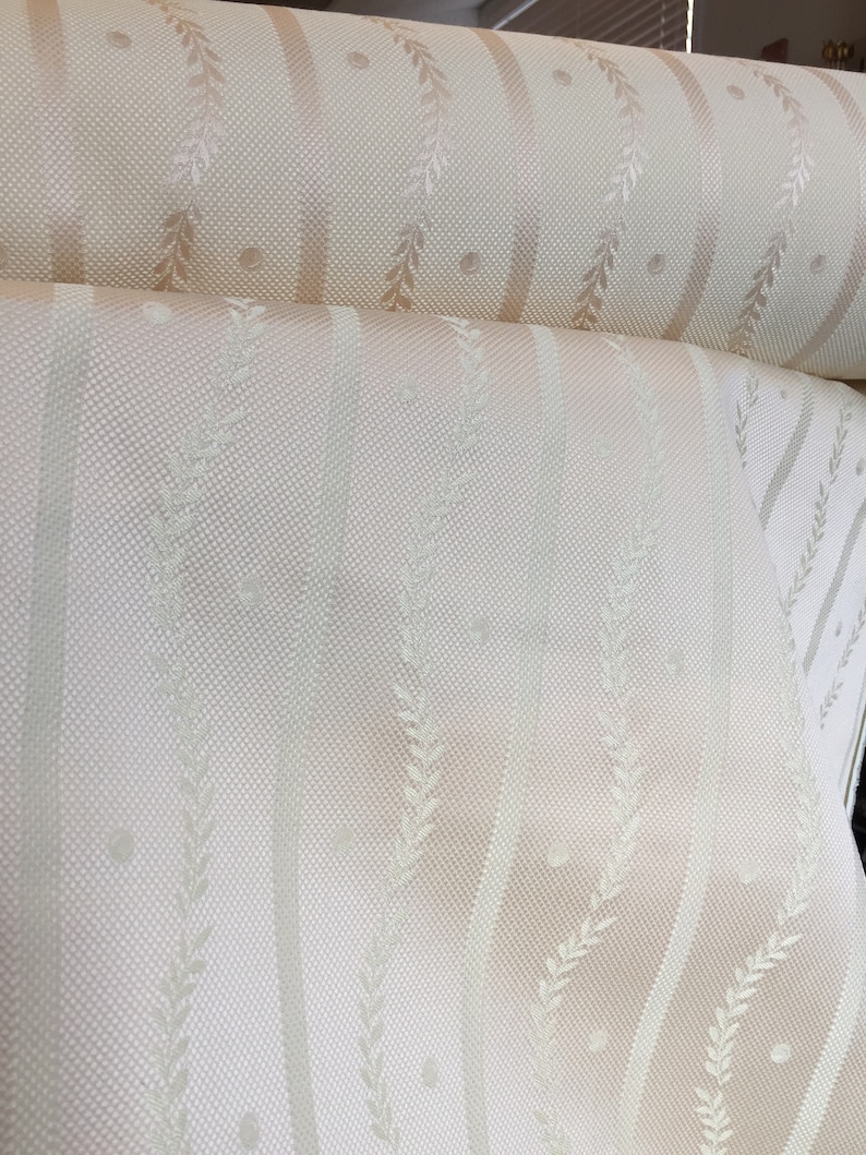 May include: Cream-colored fabric roll with a repeating pattern of vertical stripes, dotted circles, and leaf-like designs. The fabric has a subtle texture and a soft, neutral color palette, suitable for upholstery or drapery.