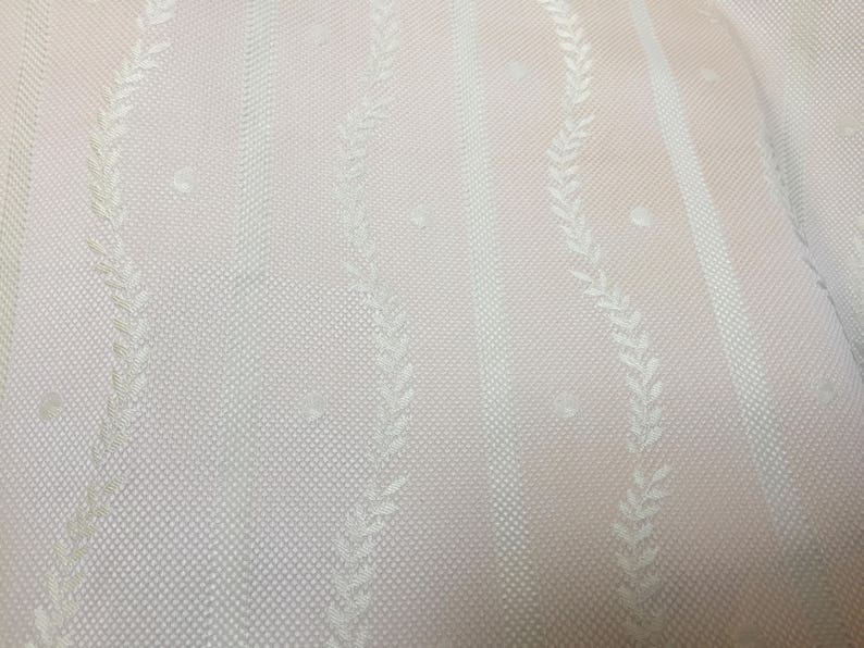 May include: Close-up of a light pink fabric with a textured, dotted pattern. Vertical stripes alternate with delicate, off-white leaf designs. The fabric has a subtle sheen, suggesting a soft, drapable material.