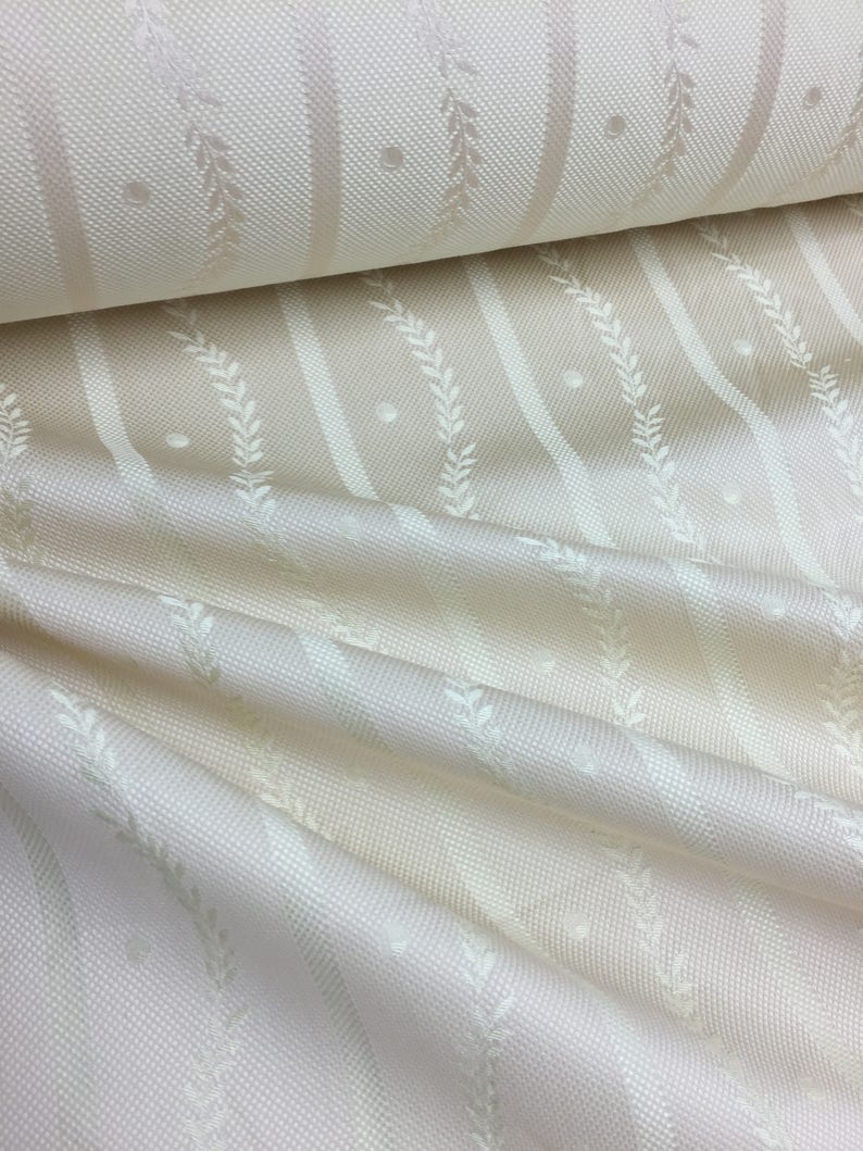 May include: Cream-colored fabric with a repeating pattern of vertical stripes, dotted accents, and stylized leaf designs. The fabric is folded, showing the texture and sheen of the material. The overall aesthetic is elegant and suitable for home decor.