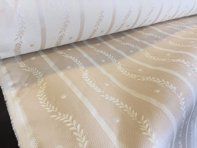 May include: A roll of fabric with a repeating pattern of vertical stripes in cream and beige. White leaf and dot details are interspersed between the stripes. The fabric has a woven texture.