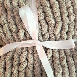 May include: A folded, chunky knit blanket in a soft, muted green-gray color. The blanket is tied with a white satin ribbon, creating a neat and gift-ready presentation. The thick knit texture is visible.