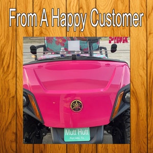 Puede incluir: Un carrito de golf Yamaha de color rosa brillante con un volante negro, espejos laterales negros y un logotipo negro de Yamaha en la parte delantera. El carrito tiene una placa de matr&iacute;cula que dice "Mutt Hutt, Port Aransas, TX".