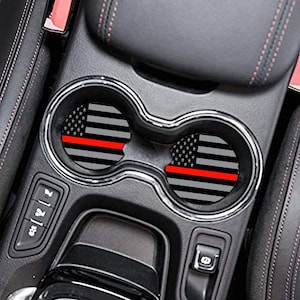 May include: Two car cup holder coasters with a design of the American flag in black, white, and gray, with a red stripe. The coasters are round and fit inside the car's cup holders. The car's interior is black and gray.
