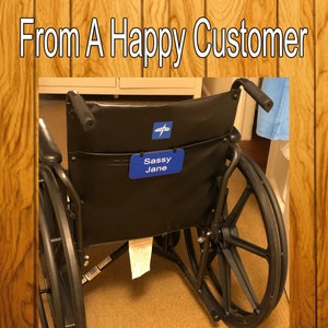 Puede incluir: Silla de ruedas negra con ruedas negras y una etiqueta de nombre azul que dice "Sassy Jane" en la parte posterior.