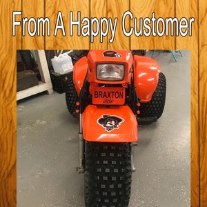 Puede incluir: ATV de tres ruedas naranja con neum&aacute;ticos negros y una placa negra que dice "BRAXTON 4539".