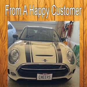 Puede incluir: Un Mini Cooper Clubman blanco con rayas de carreras negras en el cap&oacute;. El coche tiene un parachoques delantero negro y una parrilla con moldura cromada. El coche tiene una matr&iacute;cula de Ohio que dice "CHEERS".