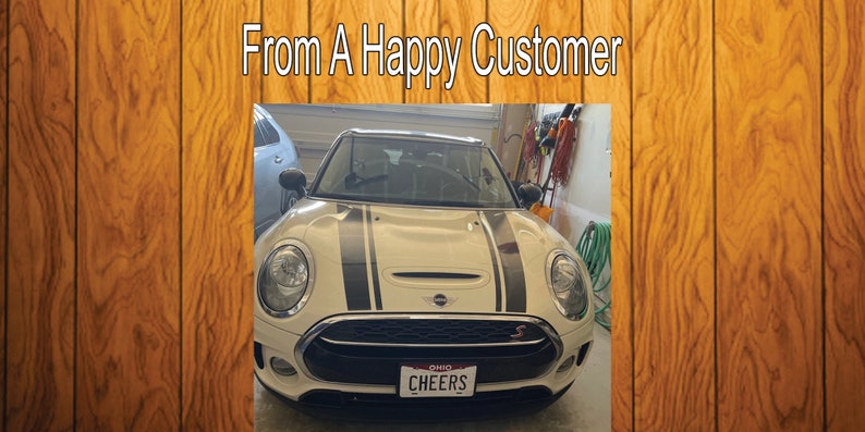 Puede incluir: Un Mini Cooper Clubman blanco con rayas de carreras negras en el cap&oacute;. El coche tiene un parachoques delantero negro y una parrilla con moldura cromada. El coche tiene una matr&iacute;cula de Ohio que dice "CHEERS".