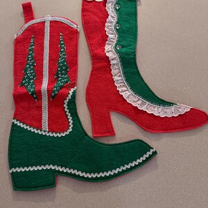May include: Two felt Christmas stockings shaped like footwear. One is a red and green cowboy boot with silver sequin tree designs. The other is a red and green boot with a white lace ruffle, eyelet trim, and clear buttons along the side.
